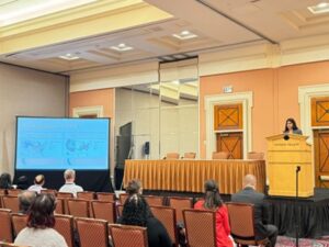 Aarti Dixit presenting at AACOM conference at podium with screen in front of seated audience