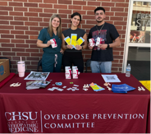 three medical students holding supplies standing behind Overdose Prevention Committee table with supplies on it