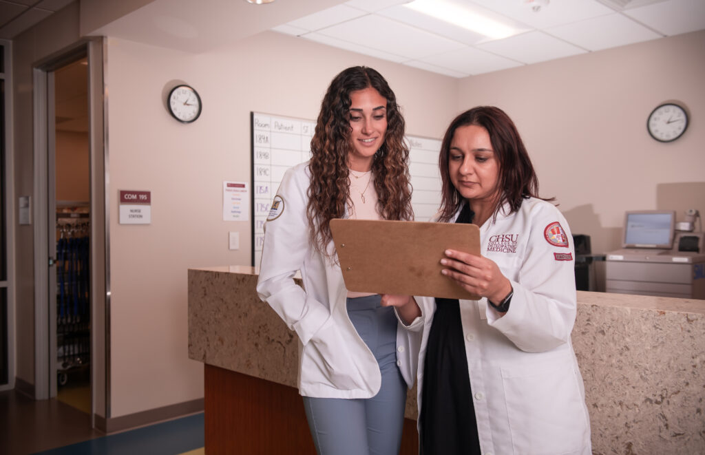 faculty in white coat reviewing chart with student in white coat at nurses station
