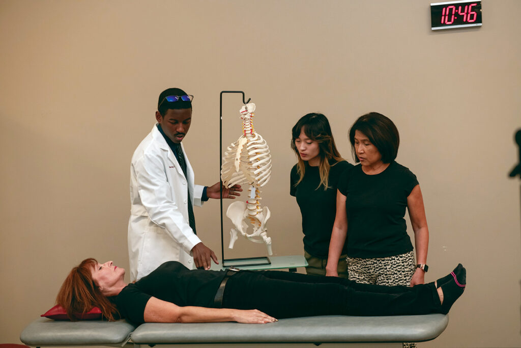 physician in white coat with skeleton model, students observing, patient laying on table