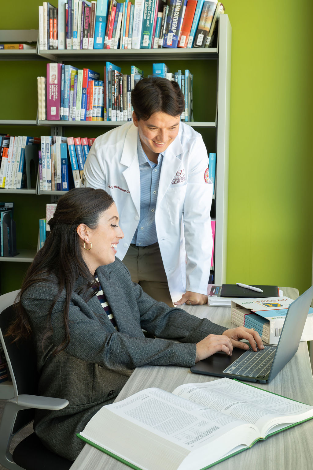 student on lap top assisting student in white coat in CHSU library