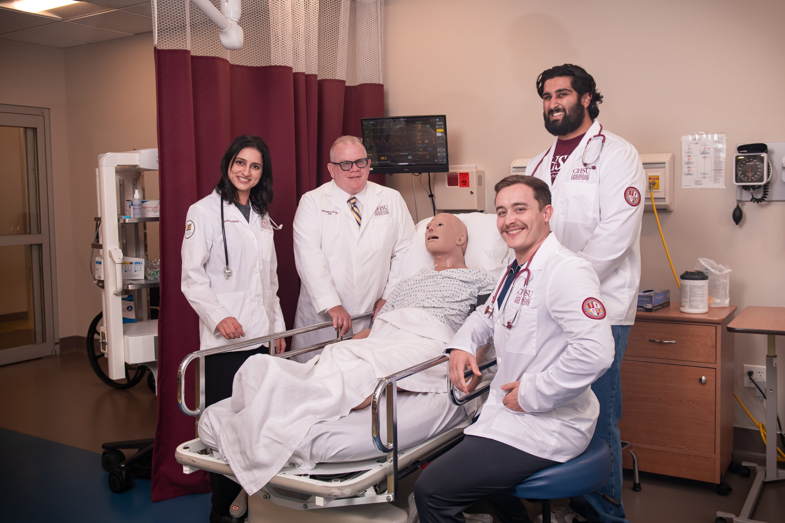 Dr. Graneto and 3 students wearing white coats examining manikin in hospital bed