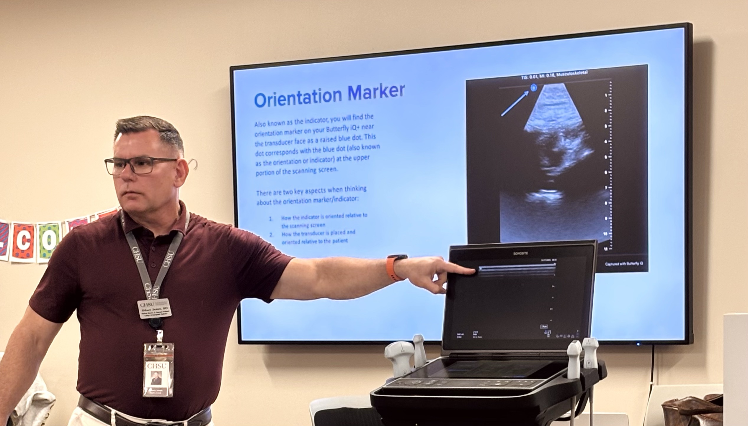 Dr. James points to an ultrasound machine during a demonstration, with a presentation slide displayed behind him.