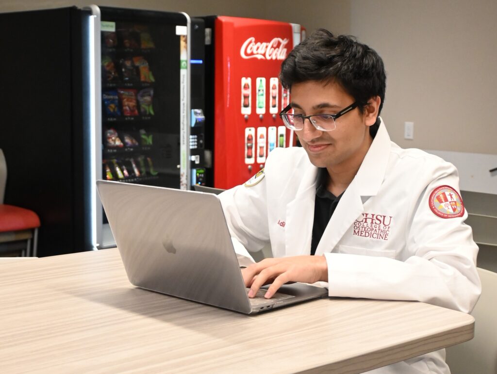 student in white coat seated at table working on laptop