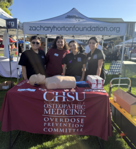 Four CHSU Overdose Prevention Committee members stand behind an outreach table with opioid overdose response training materials at SoberStock Fest.